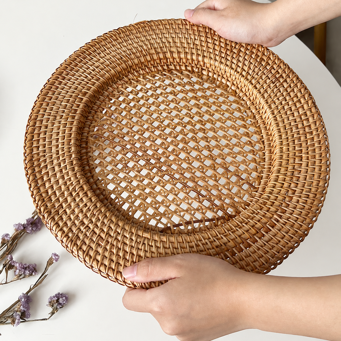 Hands holding a large eco-friendly woven rattan charger plate over white table with dried flowers