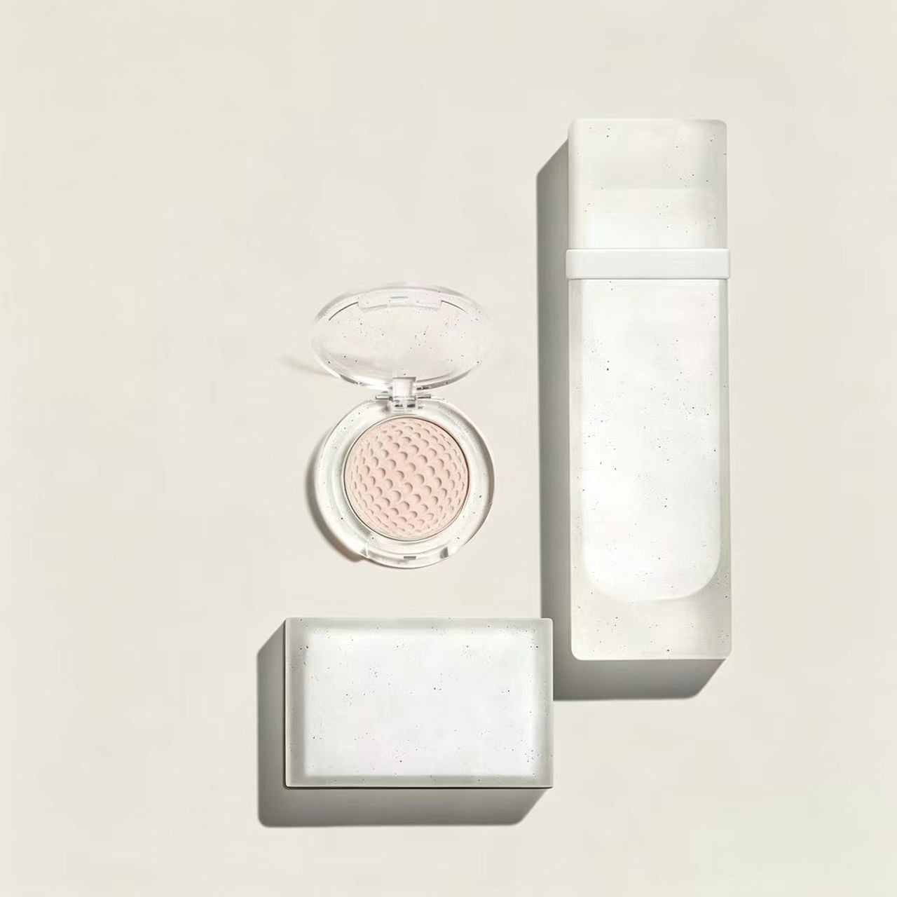Top-down view of a minimalist eco-luxury cosmetic packaging set including a refillable compact case, a frosted glass bottle, and a bar of soap.