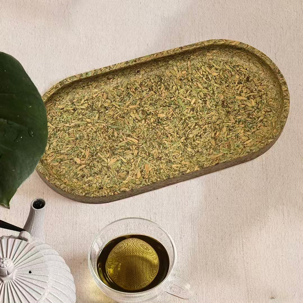 Top-down view of an organic tea leaf fiber tray alongside a ceramic teapot and transparent tea cup on linen fabric.