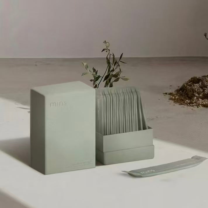A minimalist, light sage green vertical supplement box and a matching open dispenser holding stick packs, displayed on a clean organic white surface with shadow overlays and a small botanical accent.