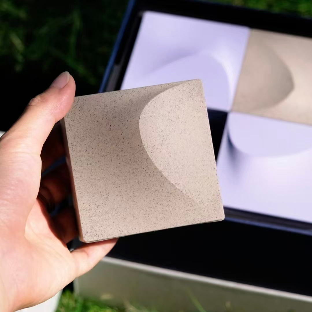 Close-up of a hand holding a minimalist geometric packaging box made from tea-fiber bio-composite on a sunny green grass background.