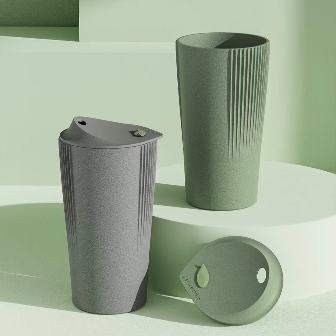  Two eco-friendly daily tumblers made from tea-fiber bio-composite in grey and olive green, featuring a speckled texture and sleek vertical grooves on a geometric platform.