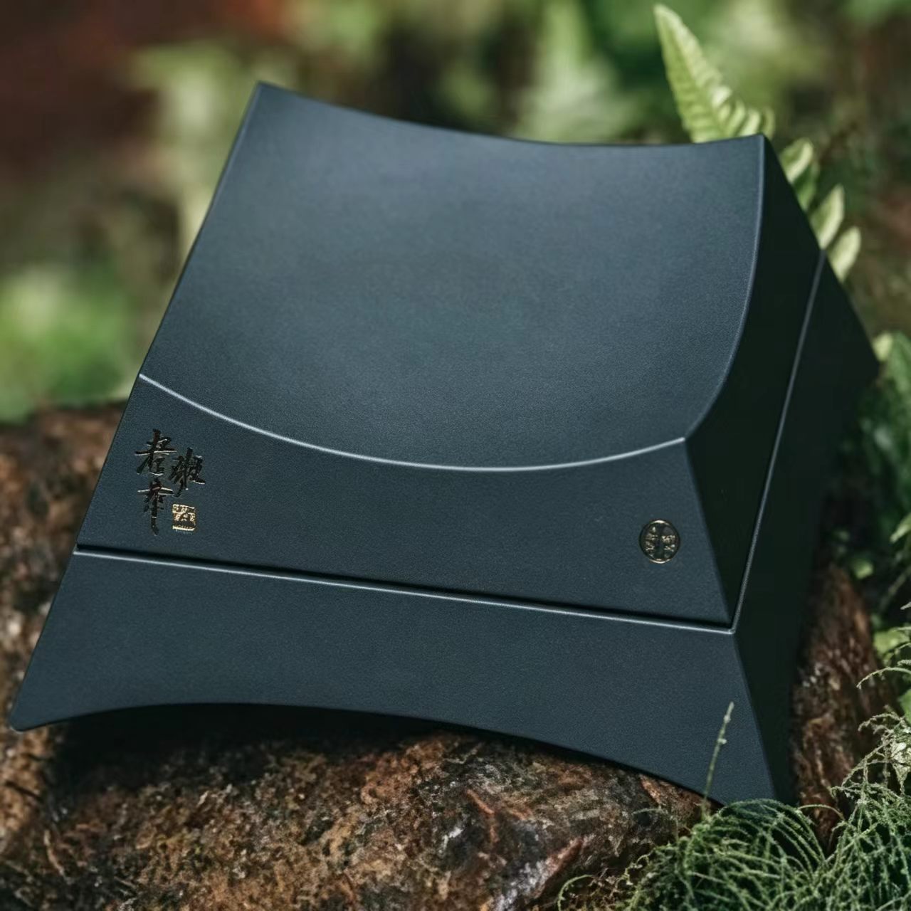 A premium dark green custom-shaped gift box with architectural concave curves, made from tea-fiber bio-composite material, displayed outdoors on organic moss.
